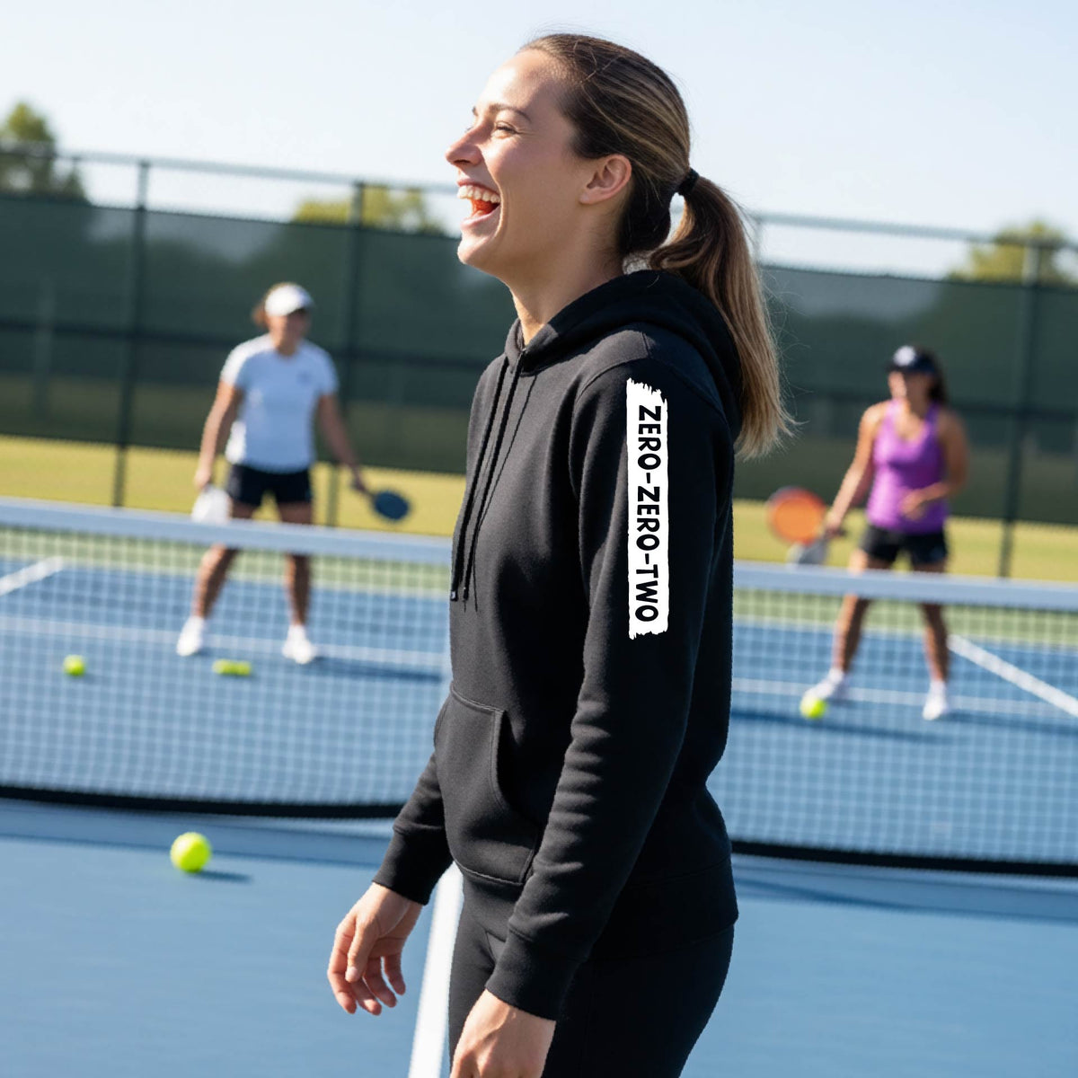 A close-up of a woman on a pickleball court wearing a black fitted hoodie pullover, with a white graphic of artistic banner with the cutout words of ‘ZERO ZERO TWO’ , on the sleeve of the hoodie. 