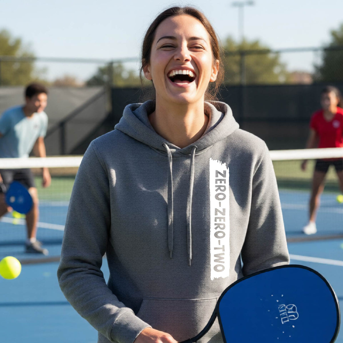 A close-up of a woman on a pickleball court wearing a gray fitted hoodie pullover, with a white graphic of artistic banner with the cutout words of ‘ZERO ZERO TWO’ , on the front of the hoodie. 