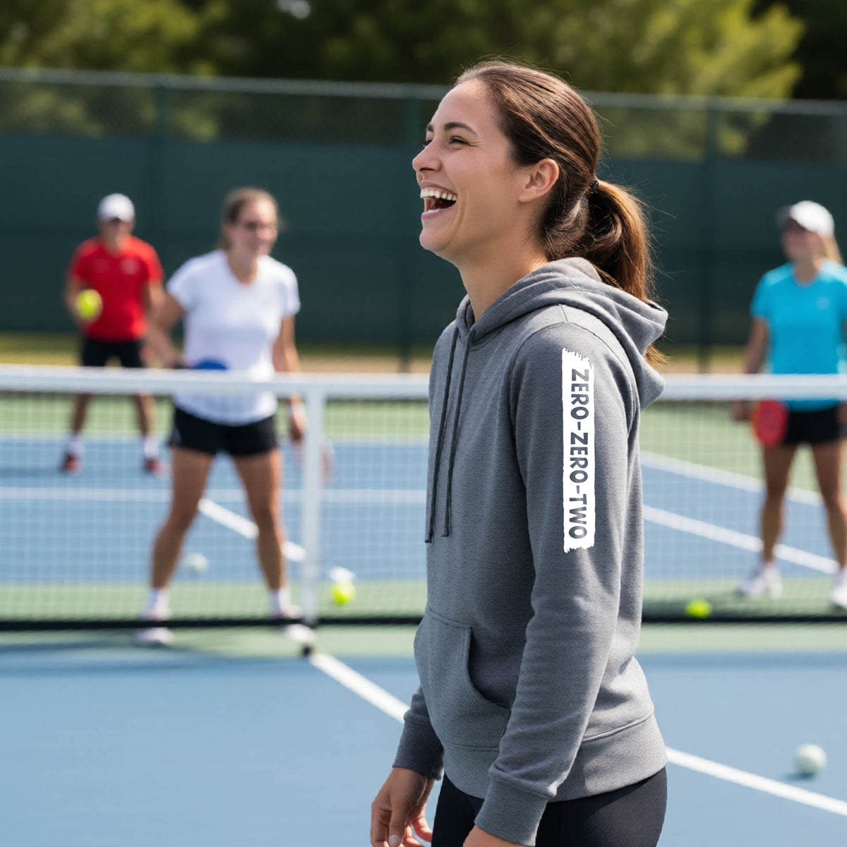 A close-up of a woman on a pickleball court wearing a gray fitted hoodie pullover, with a white graphic of artistic banner with the cutout words of ‘ZERO ZERO TWO’ , on the sleeve of the hoodie. 