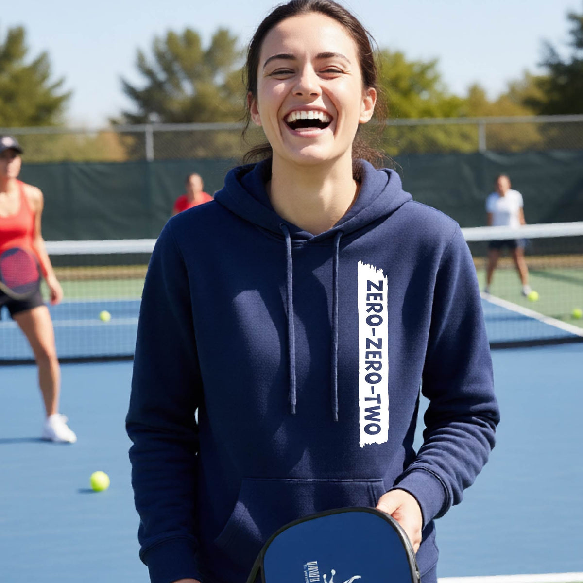 A close-up of a woman on a pickleball court wearing a navy blue fitted hoodie pullover, with a white graphic of artistic banner with the cutout words of ‘ZERO ZERO TWO’ , on the front of the hoodie. 