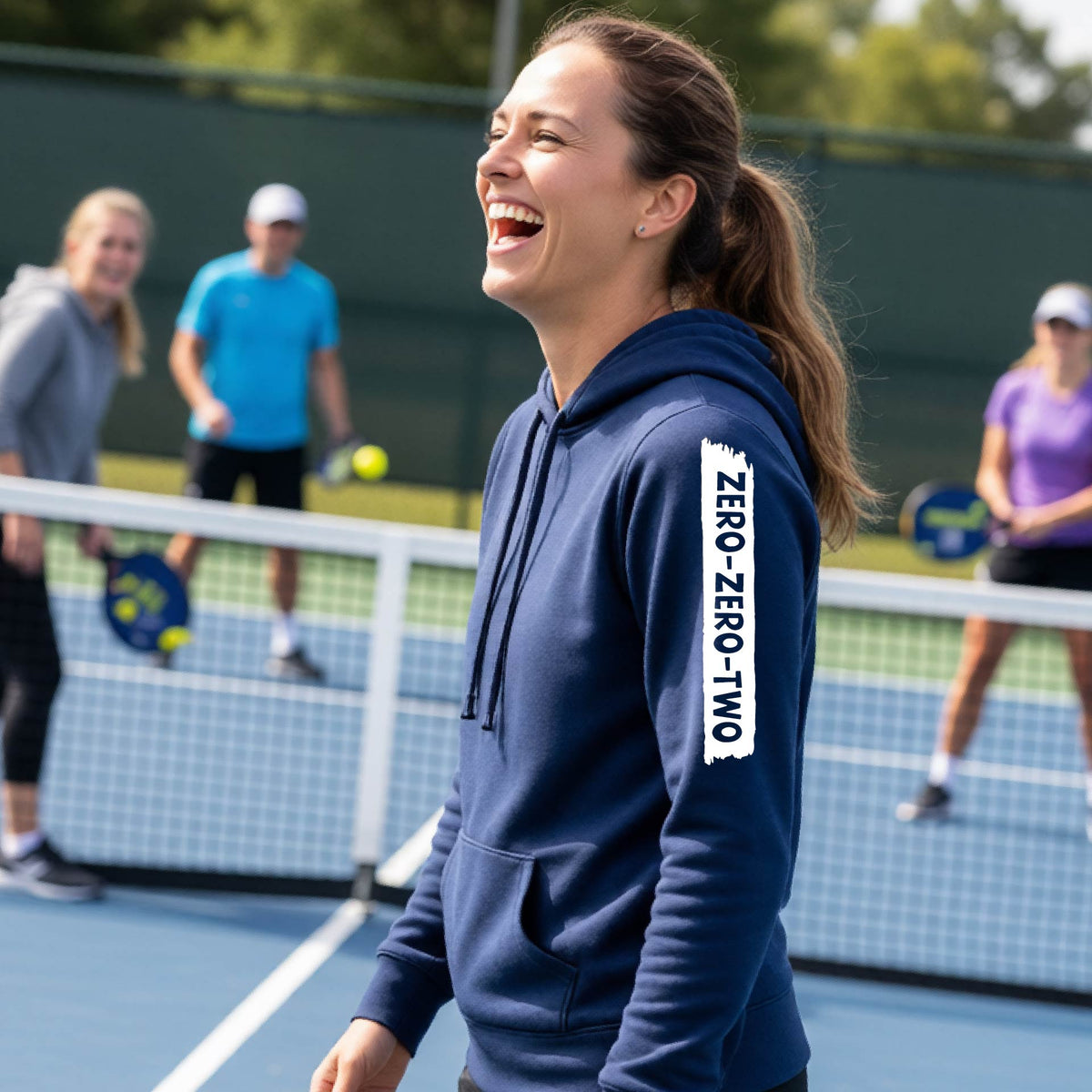A close-up of a woman on a pickleball court wearing a navy blue fitted hoodie pullover, with a white graphic of artistic banner with the cutout words of ‘ZERO ZERO TWO’ , on the sleeve of the hoodie. 