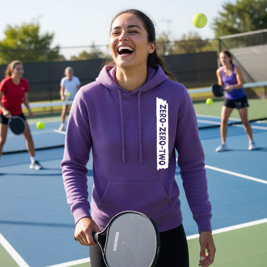 A close-up of a woman on a pickleball court wearing a purple fitted hoodie pullover, with a white graphic of artistic banner with the cutout words of ‘ZERO ZERO TWO’ , on the front of the hoodie. 