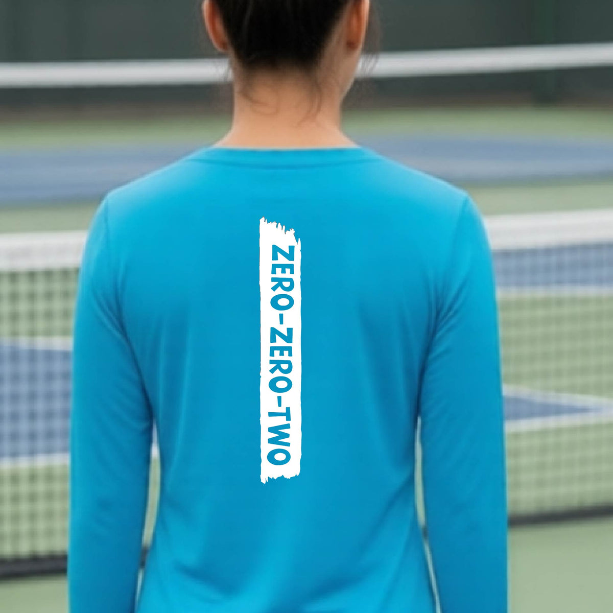 A close-up of a woman on a pickleball wearing a bright blue long-sleeve t-shirt, with a white graphic of artistic banner with the cutout words of ‘ZERO ZERO TWO’, on the back of the shirt.