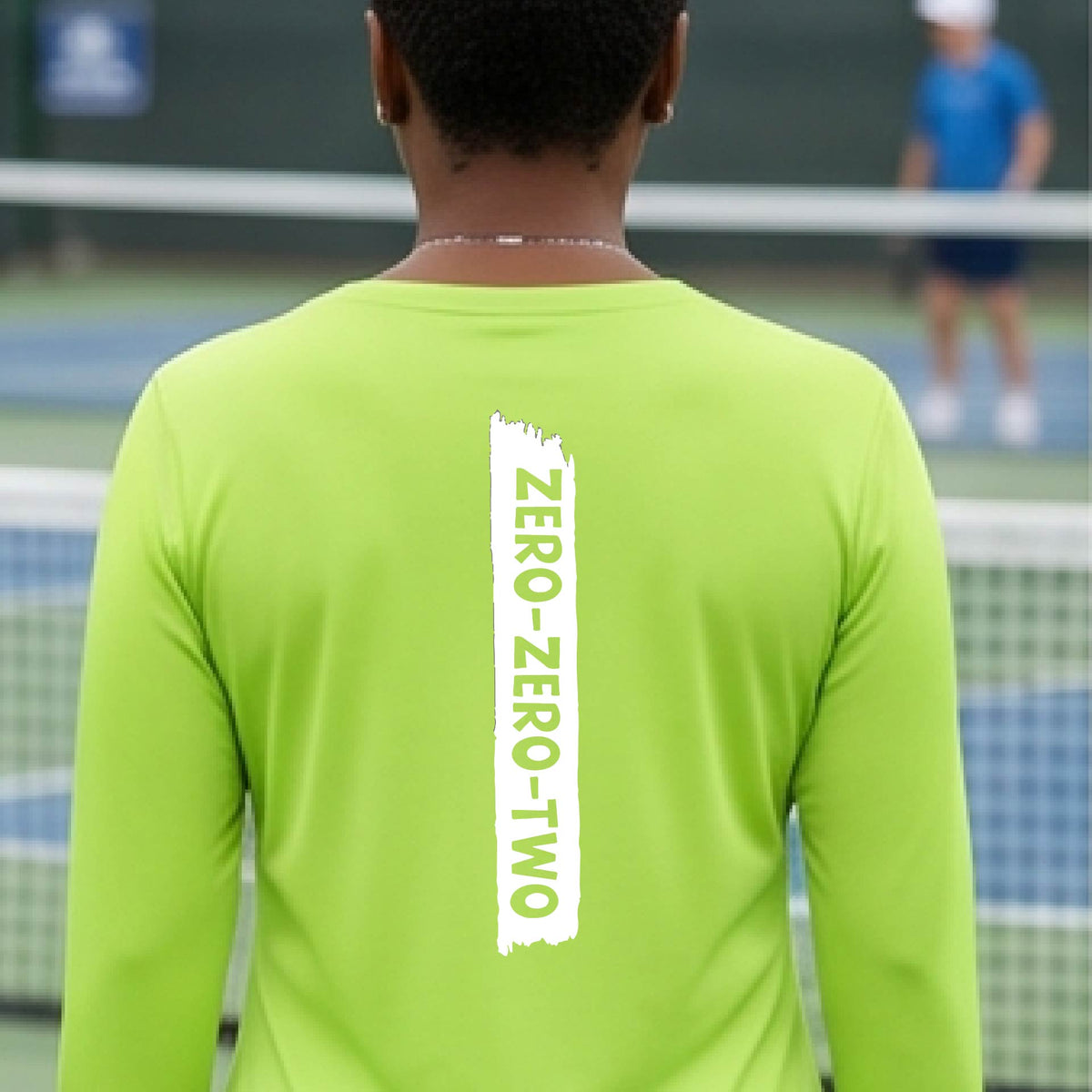 A close-up of a woman on a pickleball wearing a lime green long-sleeve t-shirt, with a white graphic of artistic banner with the cutout words of ‘ZERO ZERO TWO’, on the back of the shirt.