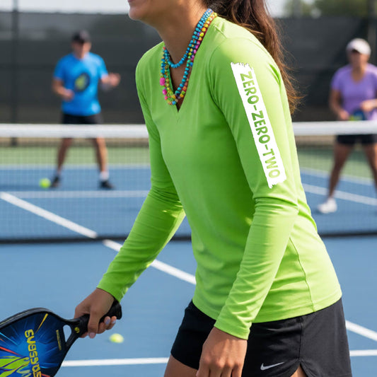 A close-up of a woman on a pickleball wearing a sleeve green long-sleeve t-shirt, with a white graphic of artistic banner with the cutout words of ‘ZERO ZERO TWO’, on the front of the shirt.