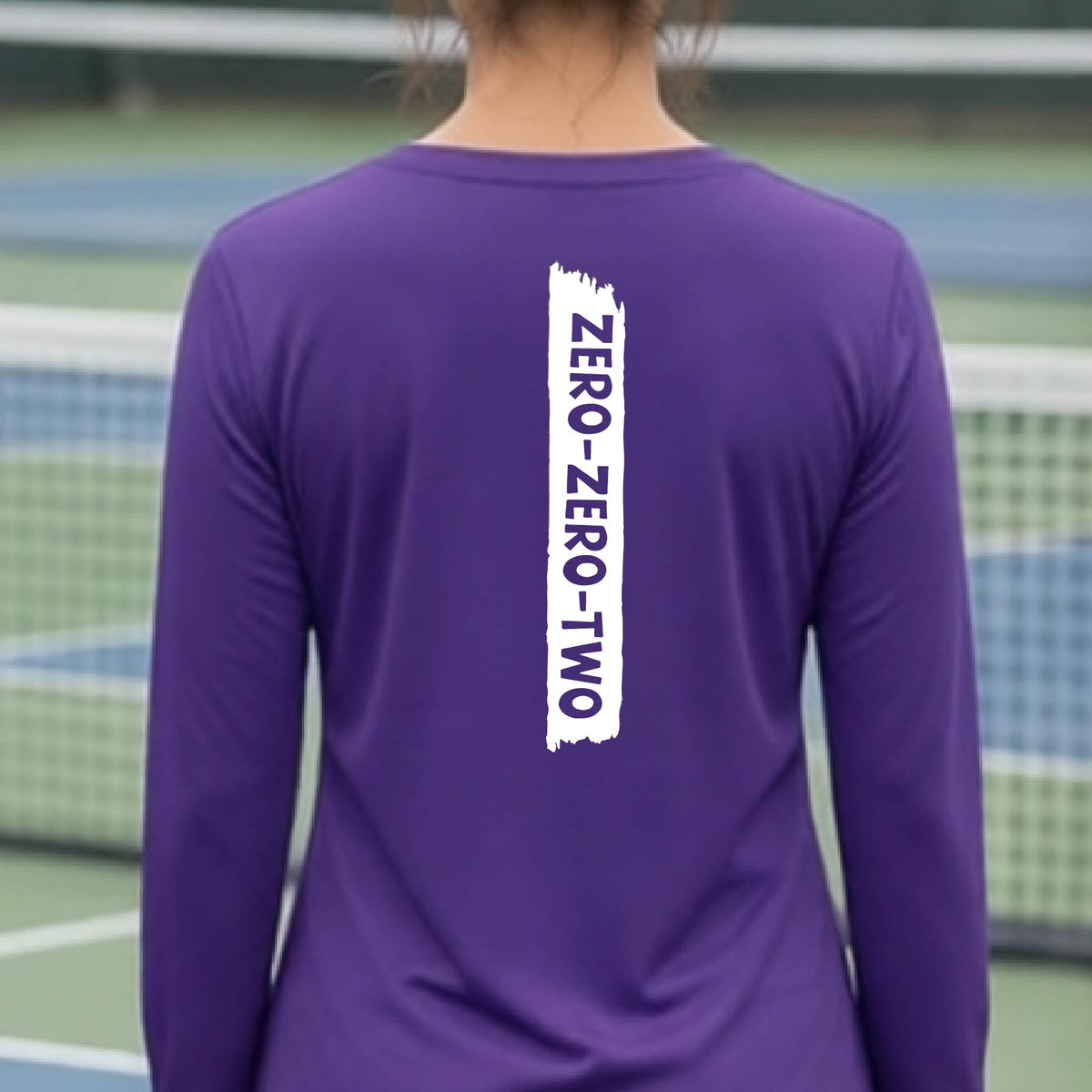 A close-up of a woman on a pickleball wearing a purple long-sleeve t-shirt, with a white graphic of artistic banner with the cutout words of ‘ZERO ZERO TWO’, on the back of the shirt.