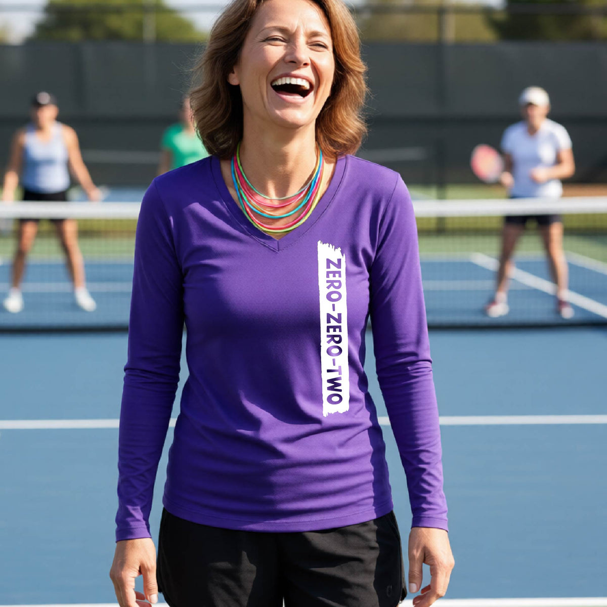 A close-up of a woman on a pickleball wearing a purple long-sleeve t-shirt, with a white graphic of artistic banner with the cutout words of ‘ZERO ZERO TWO’, on the front of the shirt.