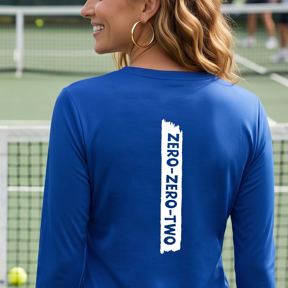 A close-up of a woman on a pickleball wearing a royal blue long-sleeve t-shirt, with a white graphic of artistic banner with the cutout words of ‘ZERO ZERO TWO’, on the back of the shirt. 