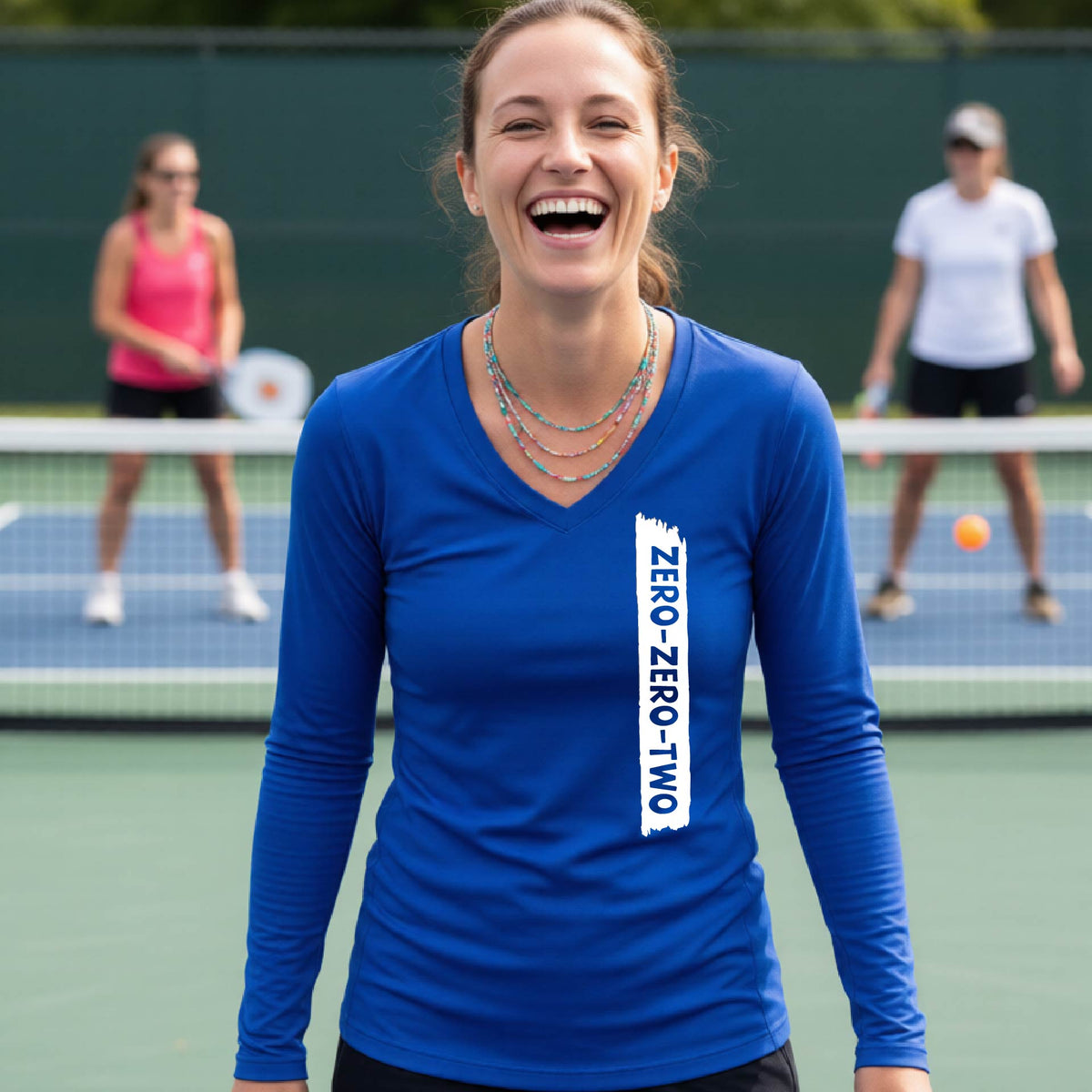 A close-up of a woman on a pickleball wearing a royal blue long-sleeve t-shirt, with a white graphic of artistic banner with the cutout words of ‘ZERO ZERO TWO’, on the front of the shirt.