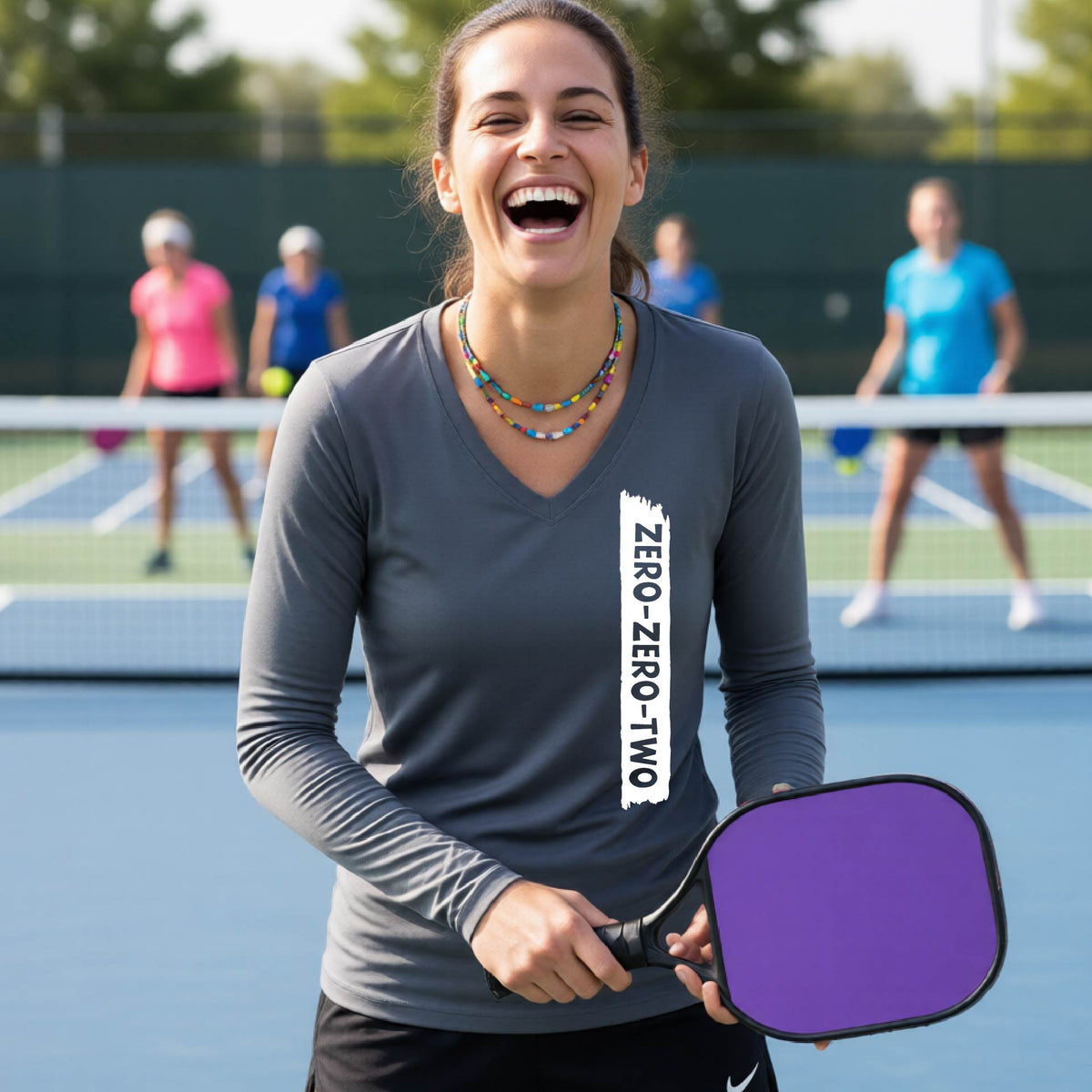 A close-up of a woman on a pickleball wearing a gray long-sleeve t-shirt, with a white graphic of artistic banner with the cutout words of ‘ZERO ZERO TWO’, on the front of the shirt.
