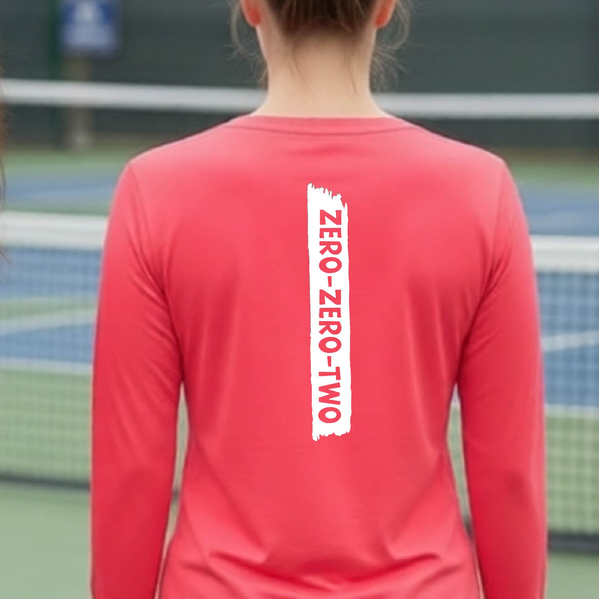 A close-up of a woman on a pickleball wearing a coral pink long-sleeve t-shirt, with a white graphic of artistic banner with the cutout words of ‘ZERO ZERO TWO’, on the back of the shirt.