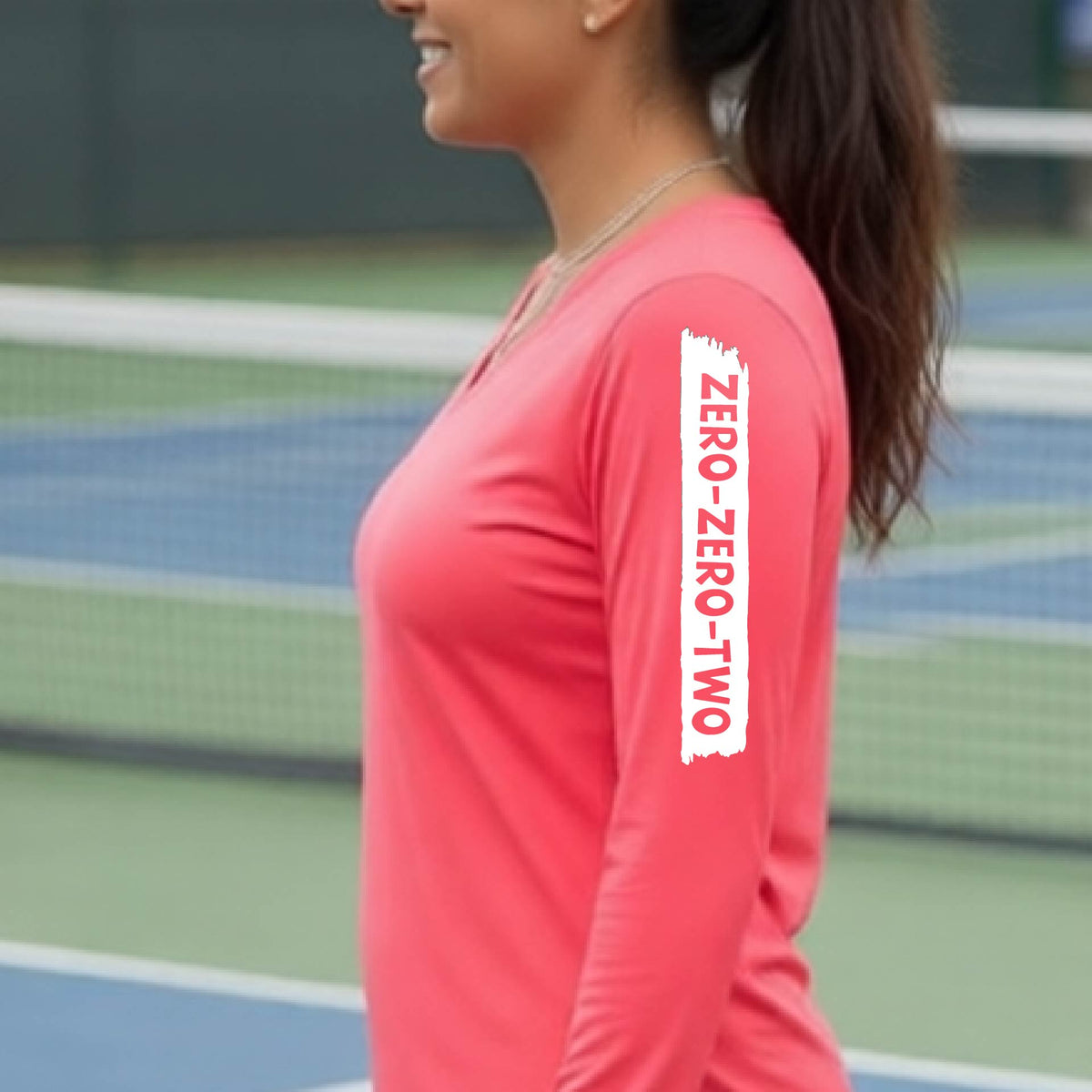 A close-up of a woman on a pickleball wearing a coral pink long-sleeve t-shirt, with a white graphic of artistic banner with the cutout words of ‘ZERO ZERO TWO’, on the sleeve of the shirt.