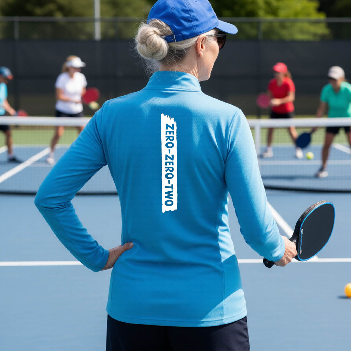 A close-up of a woman, facing backwards on a pickleball court wearing a brightl blue quarter-zip shirt,with a white graphic of artistic banner with the cutout words of ‘ZERO ZERO TWO’ , on the back of the shirt.