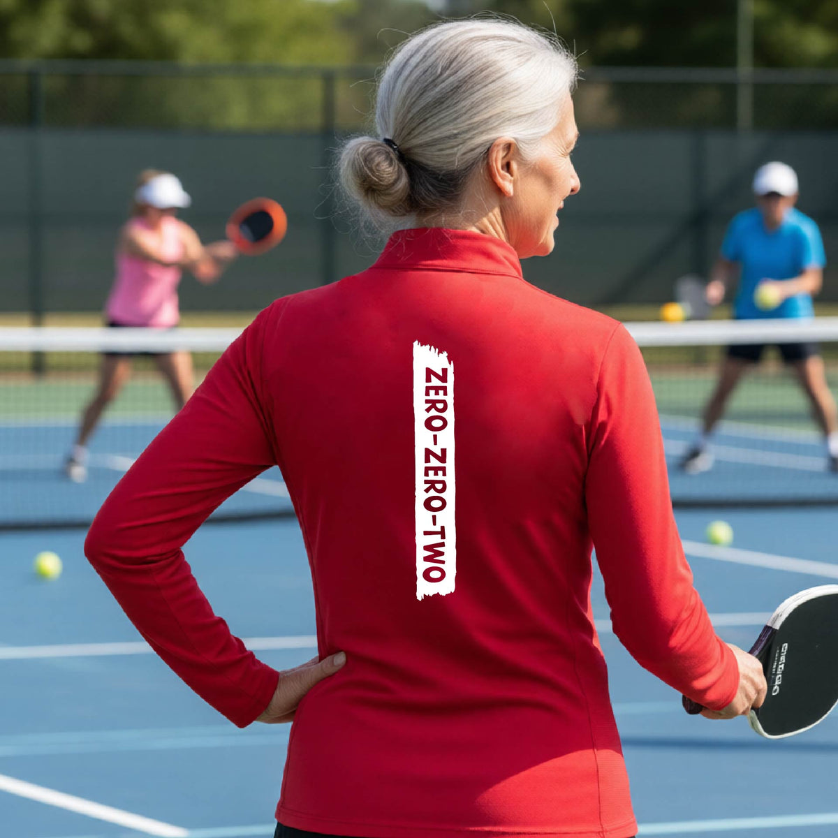 A close-up of a woman, facing backwards on a pickleball court wearing a red quarter-zip shirt,with a white graphic of artistic banner with the cutout words of ‘ZERO ZERO TWO’ , on the back of the shirt.