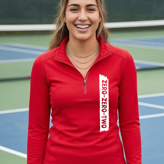 A close-up of a woman, facing backwards on a pickleball court wearing a red quarter-zip shirt,with a white graphic of artistic banner with the cutout words of ‘ZERO ZERO TWO’ , on the front of the shirt