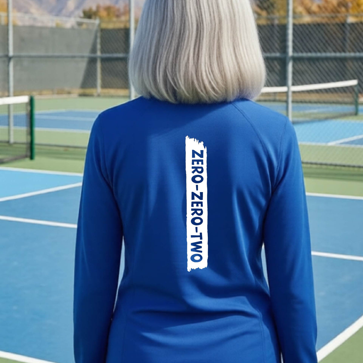 A close-up of a woman, facing backwards on a pickleball court wearing a royal blue quarter-zip shirt,with a white graphic of artistic banner with the cutout words of ‘ZERO ZERO TWO’ , on the back of the shirt.