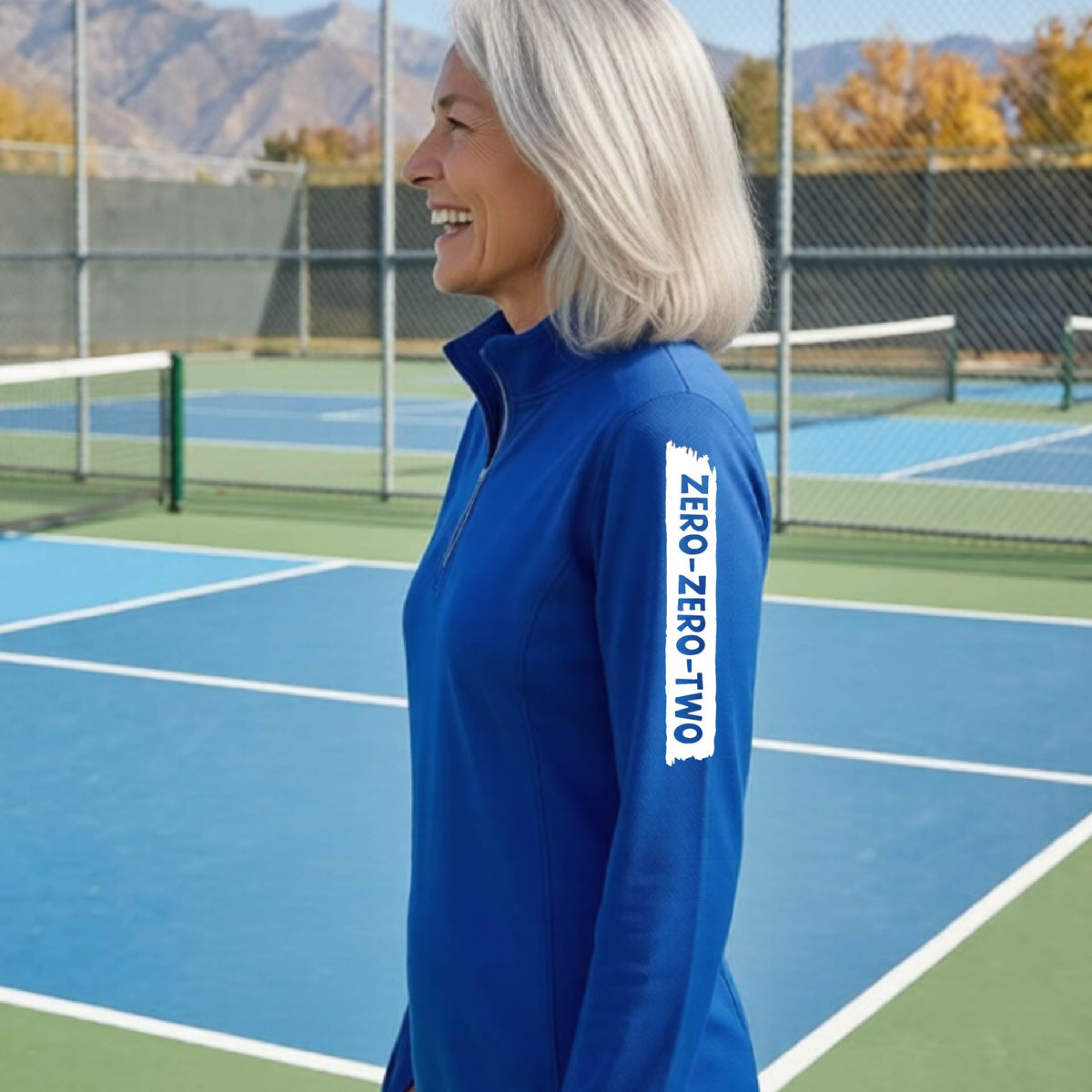 A close-up of a woman, facing backwards on a pickleball court wearing a royal blue quarter-zip shirt,with a white graphic of artistic banner with the cutout words of ‘ZERO ZERO TWO’ , on the sleeve of the shirt.