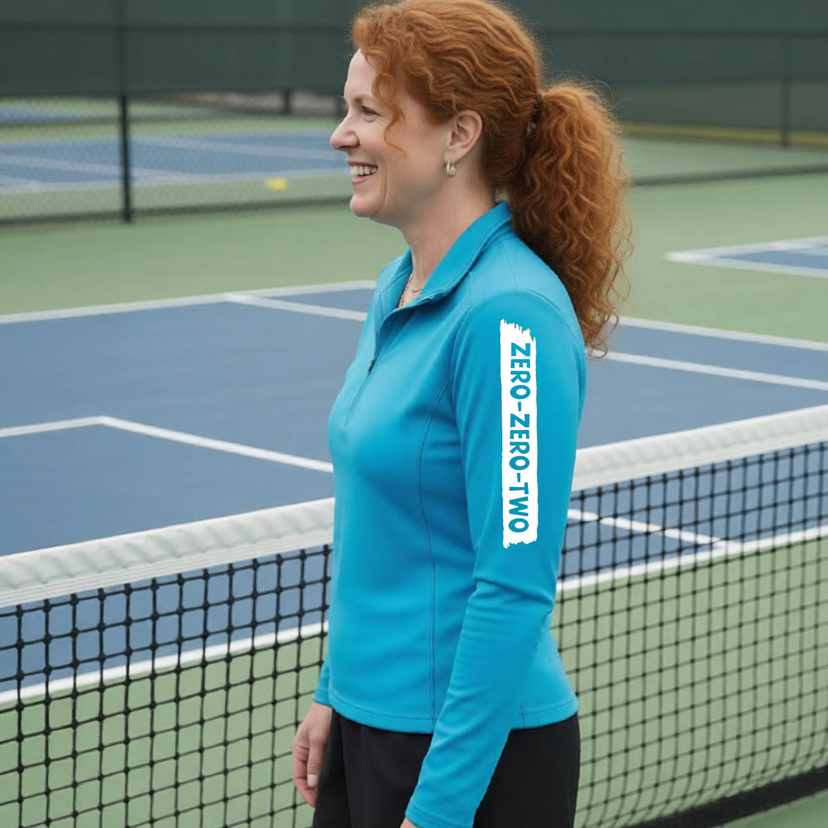 A close-up of a woman, facing backwards on a pickleball court wearing a bright blue quarter-zip shirt,with a white graphic of artistic banner with the cutout words of ‘ZERO ZERO TWO’ , on the sleeve of the shirt.