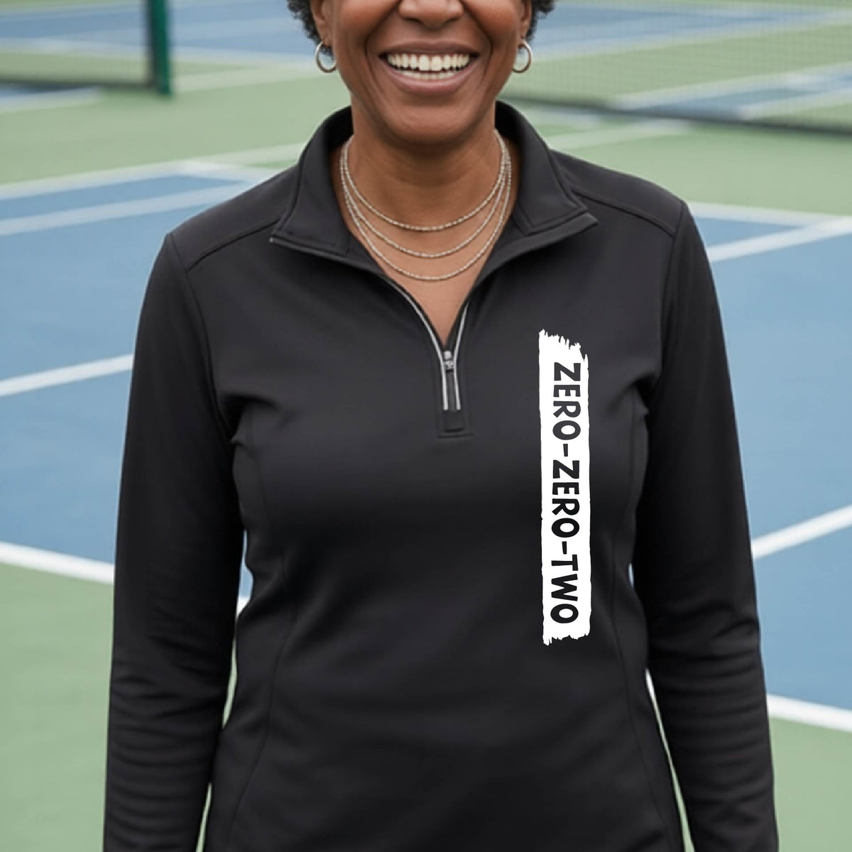 A close-up of a woman, facing backwards on a pickleball court wearing a red quarter-zip shirt,with a white graphic of artistic banner with the cutout words of ‘ZERO ZERO TWO’ , on the front of the shirt. 