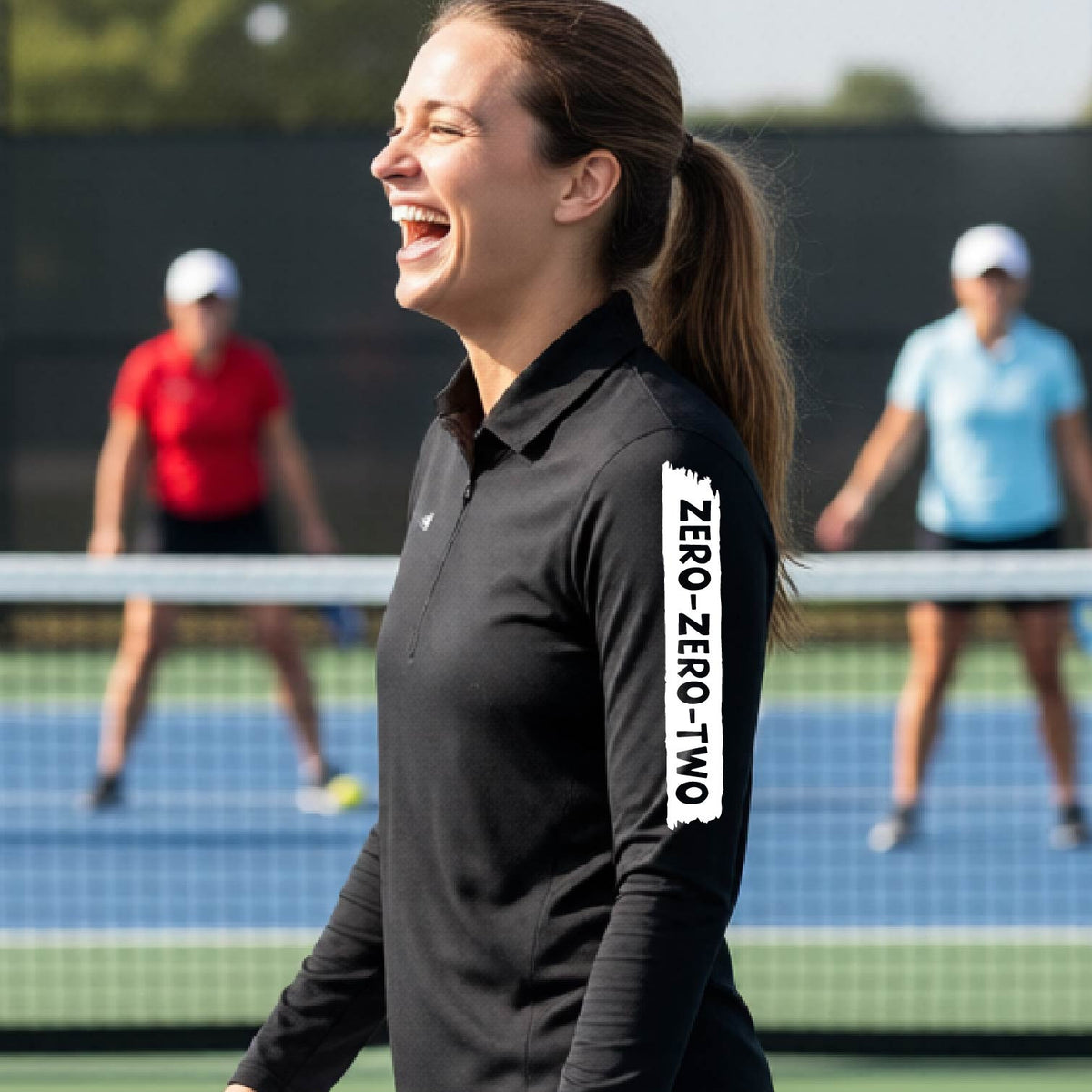 A close-up of a woman, facing backwards on a pickleball court wearing a black quarter-zip shirt,with a white graphic of artistic banner with the cutout words of ‘ZERO ZERO TWO’ , on the sleeve of the shirt.