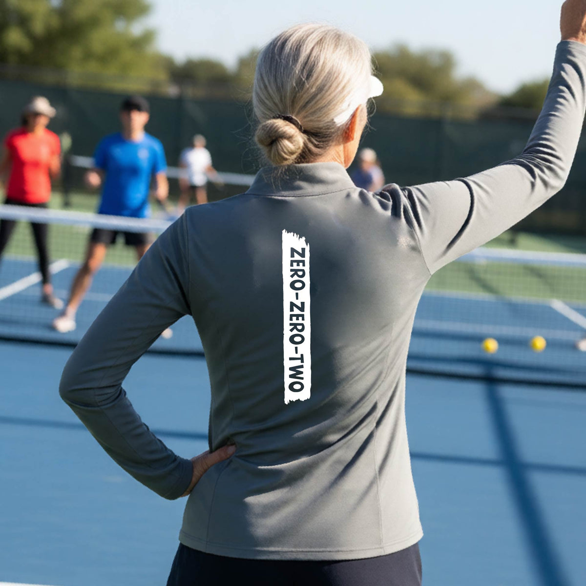 A close-up of a woman, facing backwards on a pickleball court wearing a gray quarter-zip shirt,with a white graphic of artistic banner with the cutout words of ‘ZERO ZERO TWO’ , on the back of the shirt.