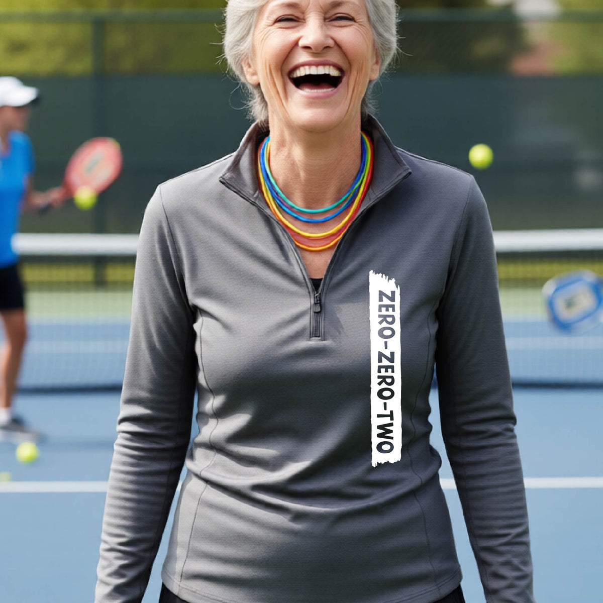 A close-up of a woman, facing backwards on a pickleball court wearing a gray quarter-zip shirt,with a white graphic of artistic banner with the cutout words of ‘ZERO ZERO TWO’ , on the front of the shirt.