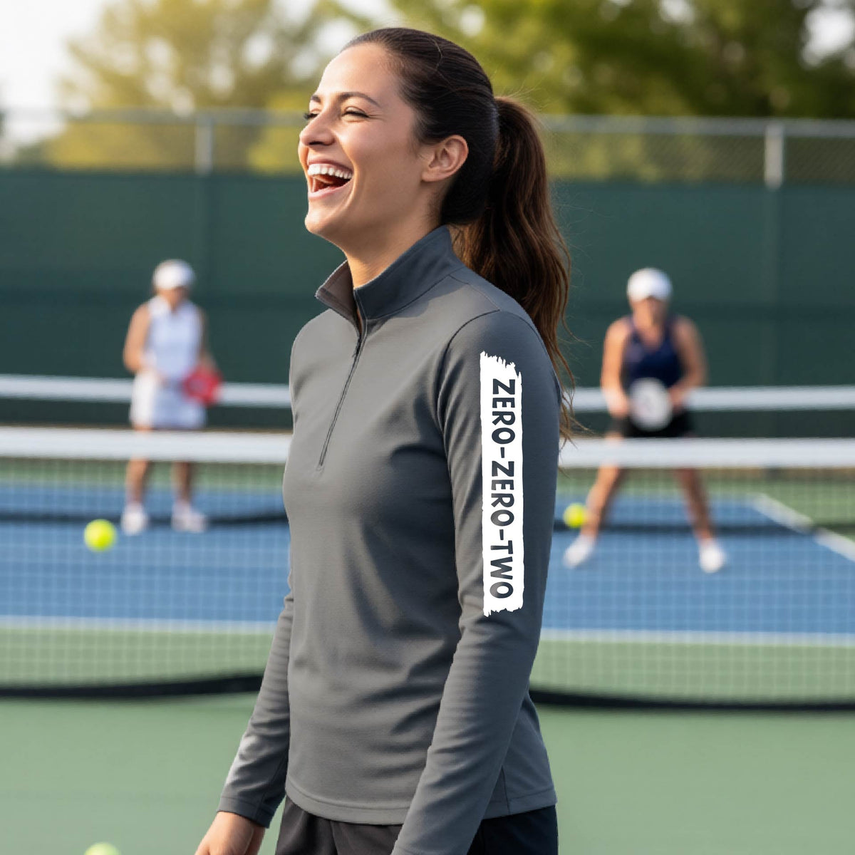 A close-up of a woman, facing backwards on a pickleball court wearing a gray quarter-zip shirt,with a white graphic of artistic banner with the cutout words of ‘ZERO ZERO TWO’ , on the sleeve of the shirt.