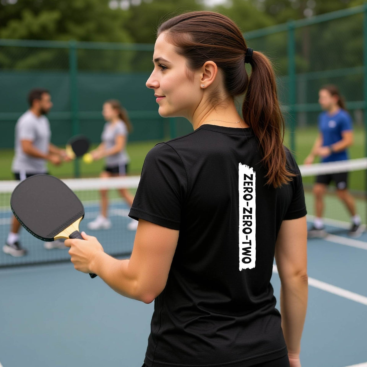 A close-up of a woman on a pickleball wearing a black short-sleeve t-shirt, with a white graphic of artistic banner with the cutout words of ‘ZERO ZERO TWO’, on the back of the shirt. 