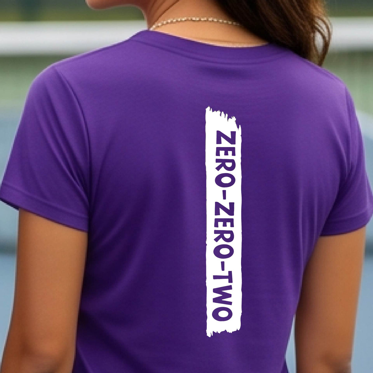 A close-up of a woman on a pickleball wearing a purple short-sleeve t-shirt, with a white graphic of artistic banner with the cutout words of ‘ZERO ZERO TWO’, on the back of the shirt. 