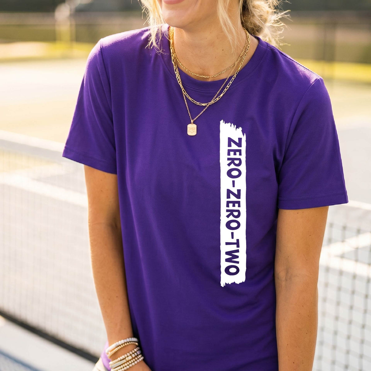 A close-up of a woman on a pickleball wearing a purple short-sleeve t-shirt, with a white graphic of artistic banner with the cutout words of ‘ZERO ZERO TWO’, on the front of the shirt. 