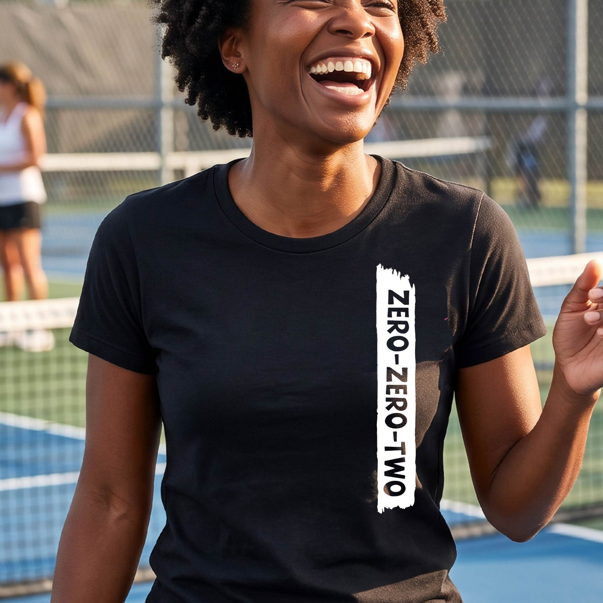 A close-up of a woman on a pickleball wearing a black short-sleeve t-shirt, with a white graphic of artistic banner with the cutout words of ‘ZERO ZERO TWO’, on the front of the shirt. 
