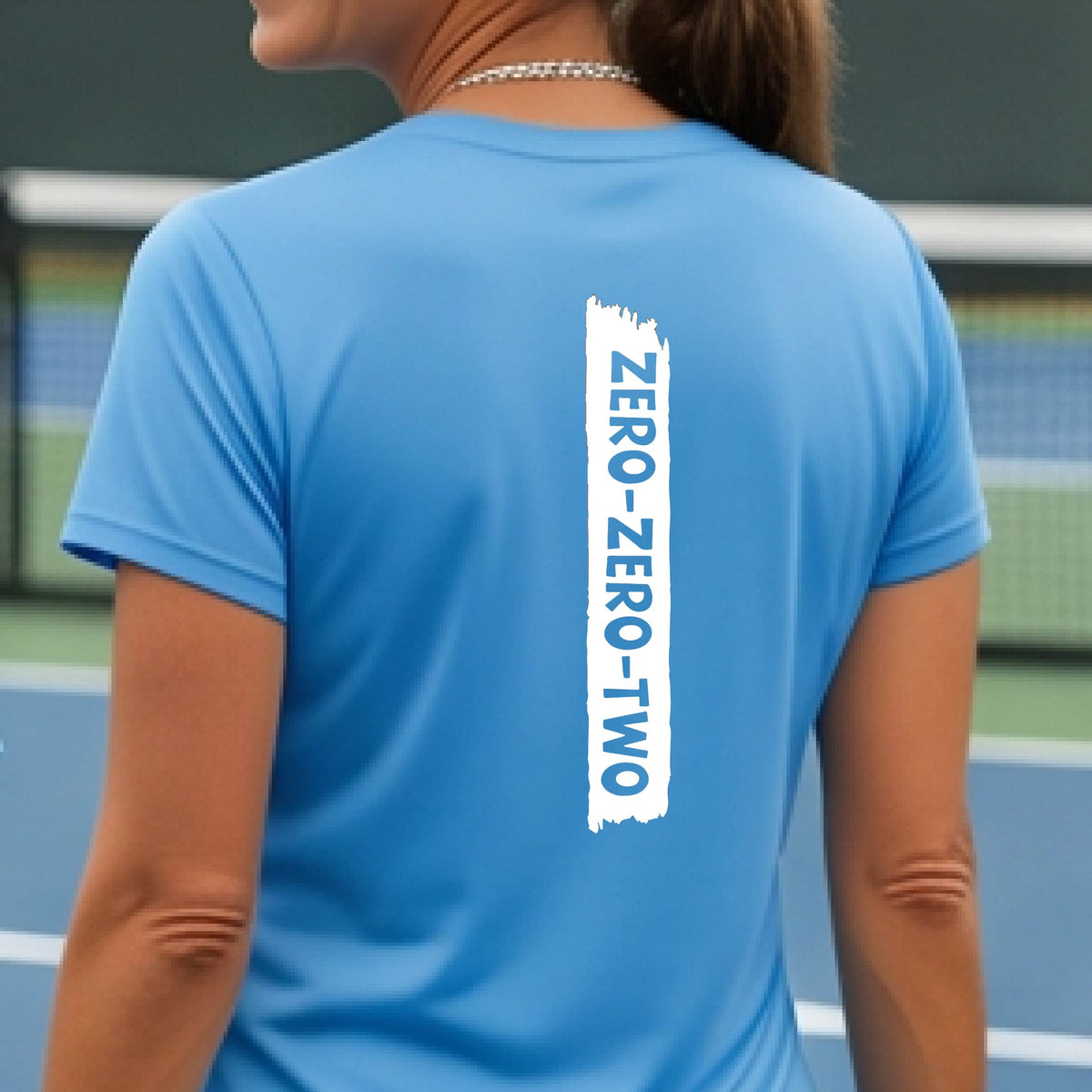 A close-up of a woman on a pickleball wearing a blue short-sleeve t-shirt, with a white graphic of artistic banner with the cutout words of ‘ZERO ZERO TWO’, on the back of the shirt.