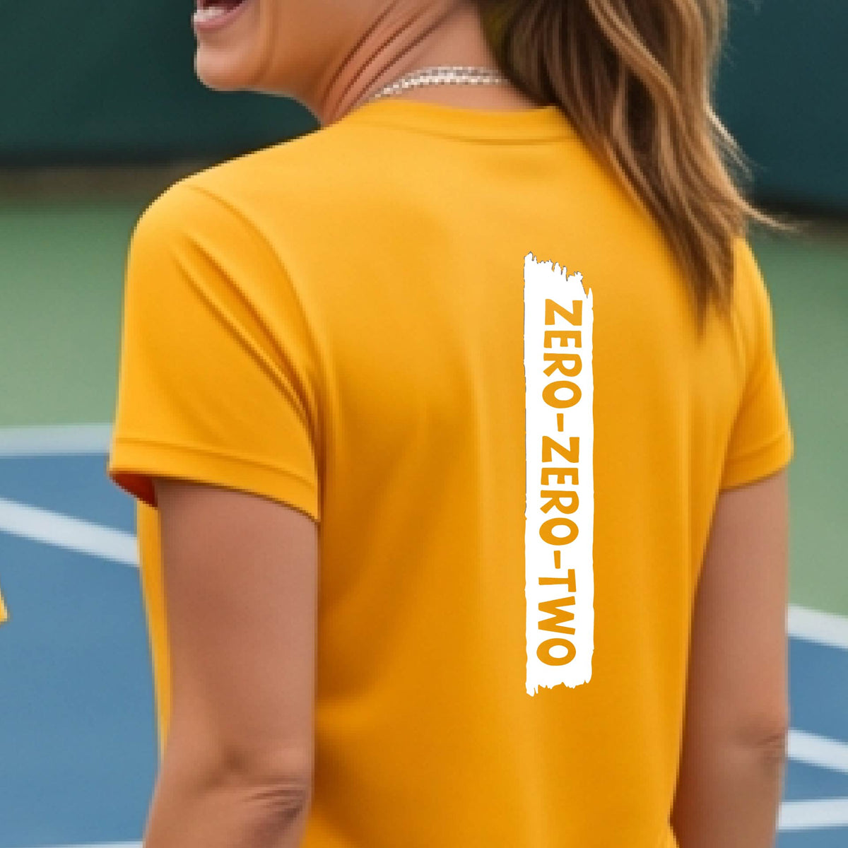 A close-up of a woman on a pickleball wearing a mustard yellow gold short-sleeve t-shirt, with a white graphic of artistic banner with the cutout words of ‘ZERO ZERO TWO’, on the back of the shirt. 