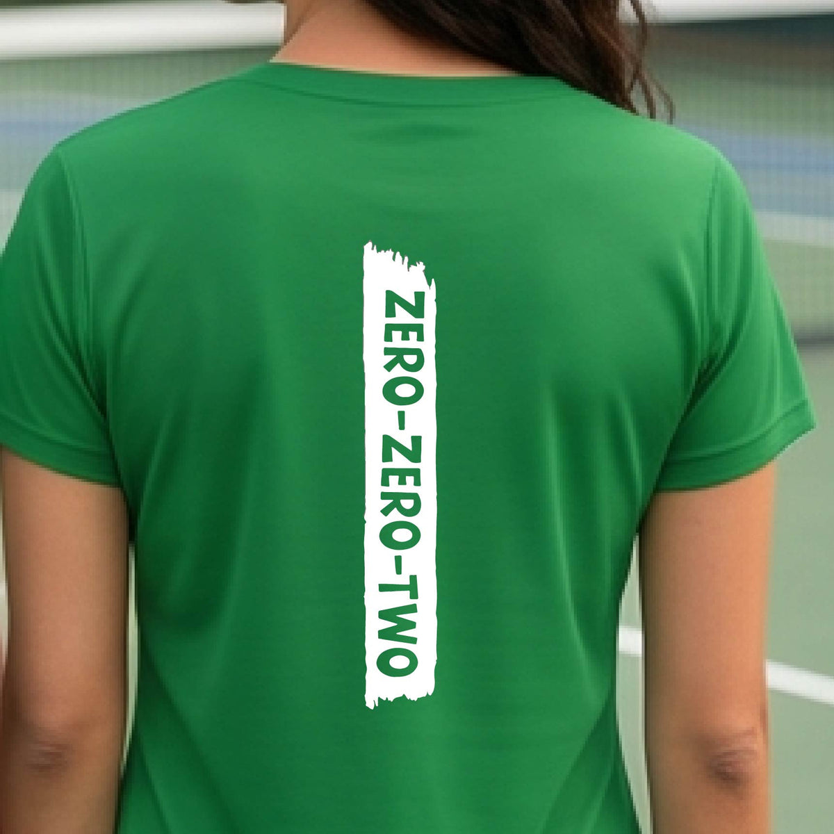 A close-up of a woman on a pickleball wearing a green short-sleeve t-shirt, with a white graphic of artistic banner with the cutout words of ‘ZERO ZERO TWO’, on the back of the shirt. 