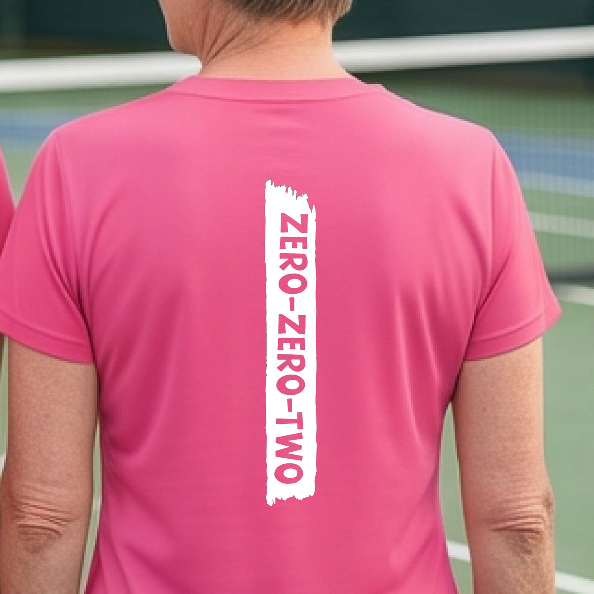 A close-up of a woman on a pickleball wearing a pink short-sleeve t-shirt, with a white graphic of artistic banner with the cutout words of ‘ZERO ZERO TWO’, on the back of the shirt. 