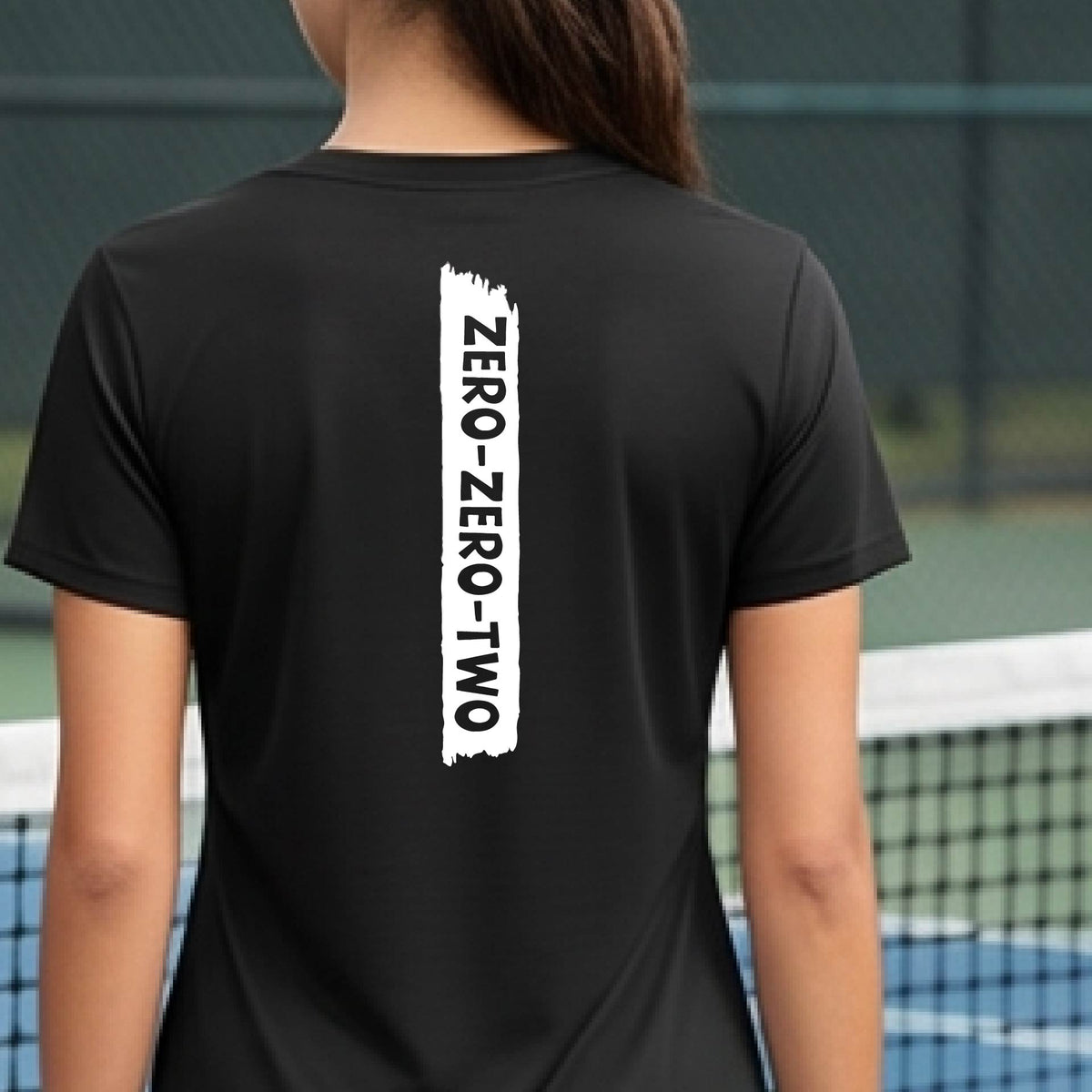 A close-up of a woman on a pickleball wearing a black short-sleeve v-neck t-shirt, with a white graphic of artistic banner with the cutout words of ‘ZERO ZERO TWO’, on the back of the shirt