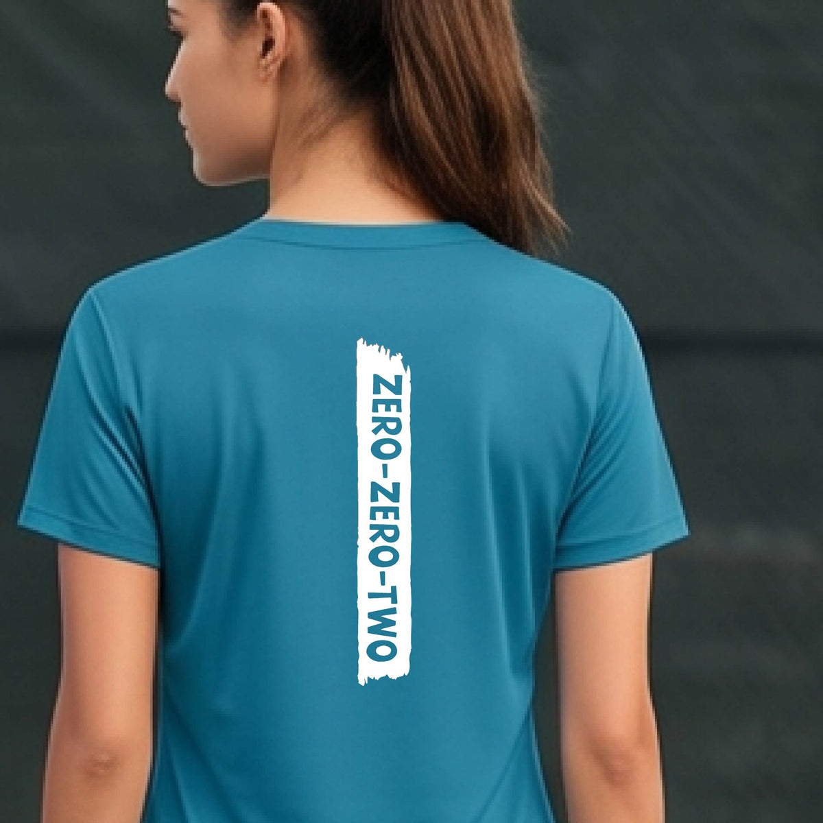 A close-up of a woman on a pickleball wearing a teal short-sleeve v-neck t-shirt, with a white graphic of artistic banner with the cutout words of ‘ZERO ZERO TWO’, on the back of the shirt.