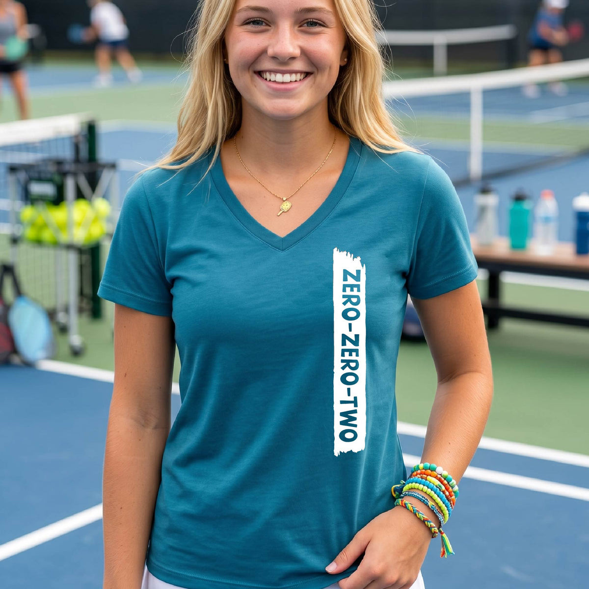 A close-up of a woman on a pickleball wearing a teal short-sleeve v-neck t-shirt, with a white graphic of artistic banner with the cutout words of ‘ZERO ZERO TWO’, on the front of the shirt.