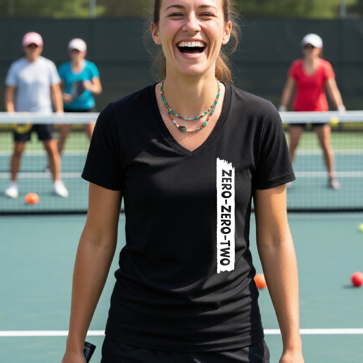 A close-up of a woman on a pickleball wearing a black short-sleeve v-neck t-shirt, with a white graphic of artistic banner with the cutout words of ‘ZERO ZERO TWO’, on the front of the shirt.