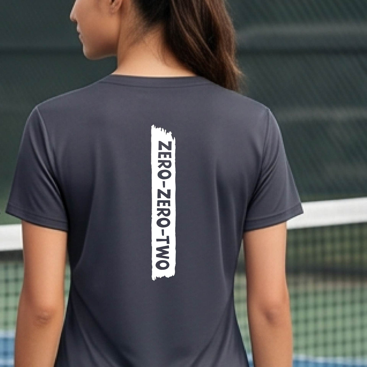A close-up of a woman on a pickleball wearing a gray short-sleeve v-neck t-shirt, with a white graphic of artistic banner with the cutout words of ‘ZERO ZERO TWO’, on the back of the shirt