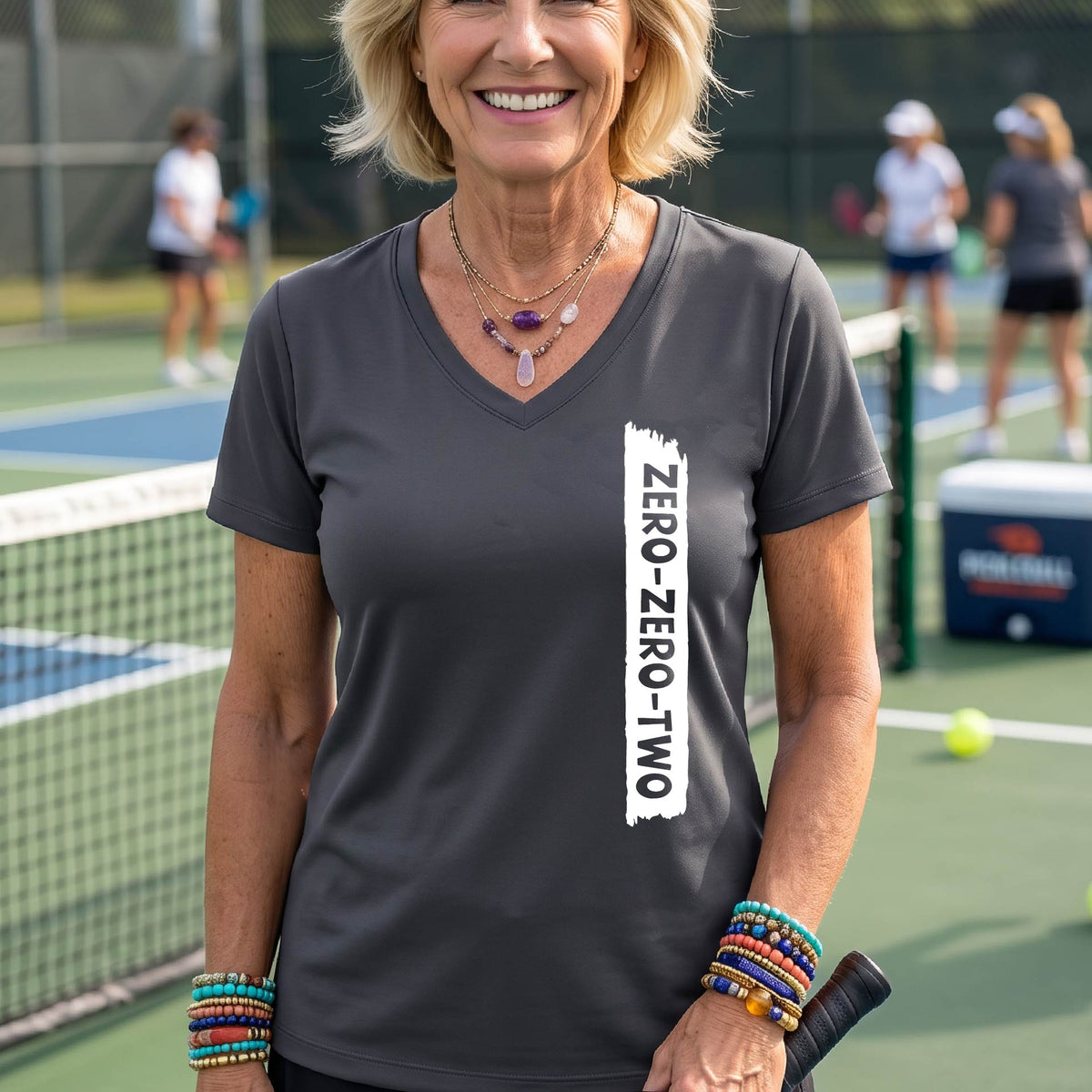 A close-up of a woman on a pickleball wearing a gray short-sleeve v-neck t-shirt, with a white graphic of artistic banner with the cutout words of ‘ZERO ZERO TWO’, on the front of the shirt