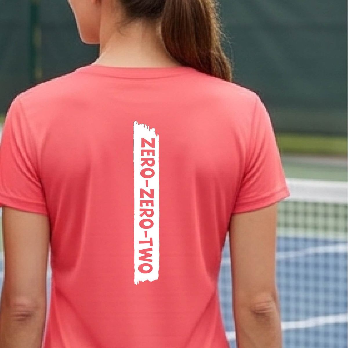 A close-up of a woman on a pickleball wearing a coral pink short-sleeve v-neck t-shirt, with a white graphic of artistic banner with the cutout words of ‘ZERO ZERO TWO’, on the back of the shirt