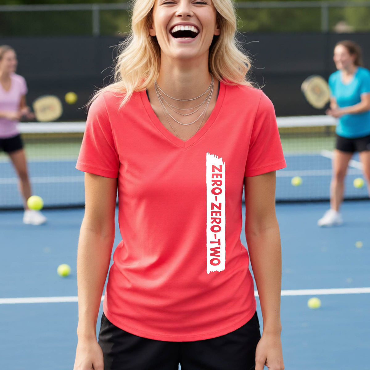 A close-up of a woman on a pickleball wearing a coral pink short-sleeve v-neck t-shirt, with a white graphic of artistic banner with the cutout words of ‘ZERO ZERO TWO’, on the front of the shirt.