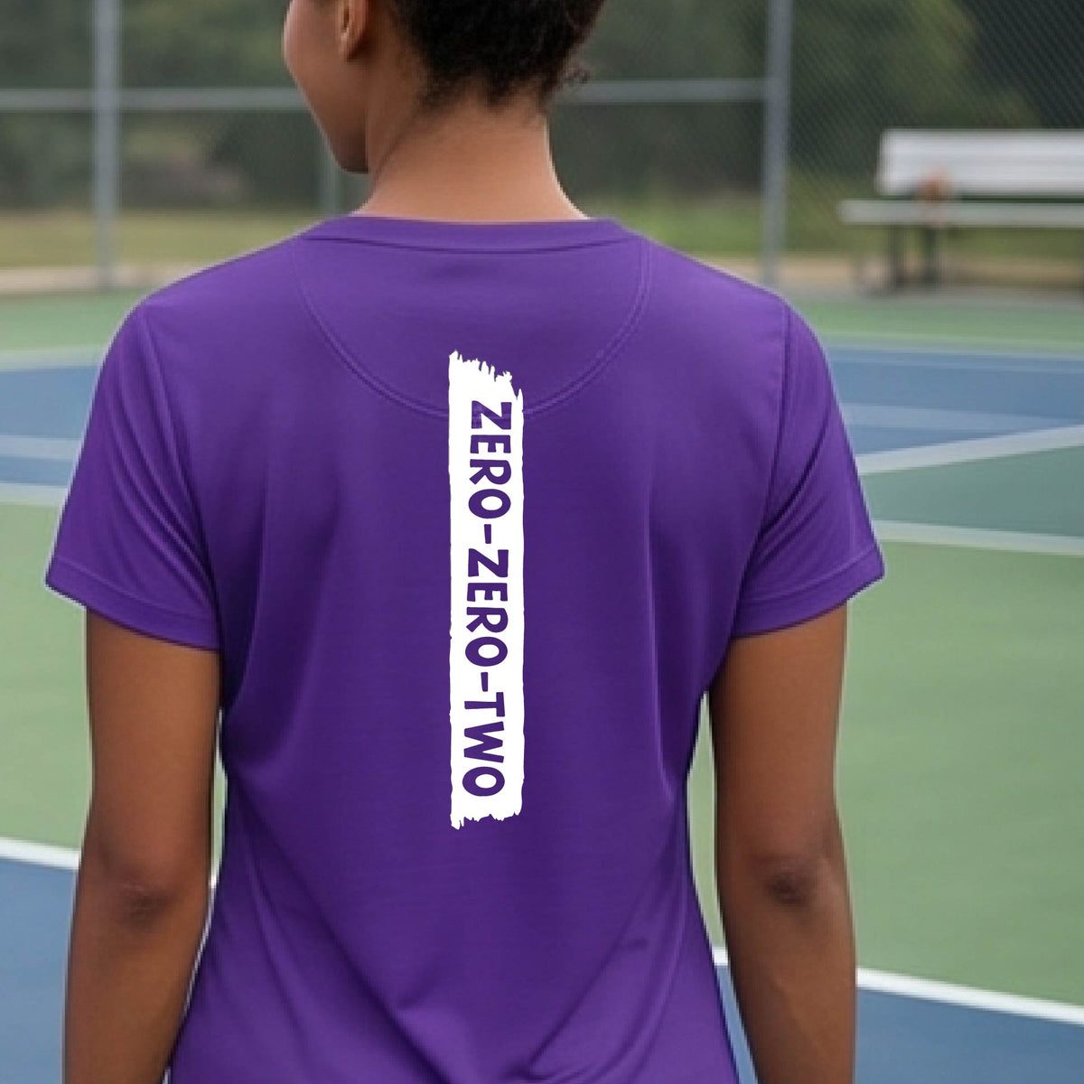 A close-up of a woman on a pickleball wearing a purple short-sleeve v-neck t-shirt, with a white graphic of artistic banner with the cutout words of ‘ZERO ZERO TWO’, on the back of the shirt. 