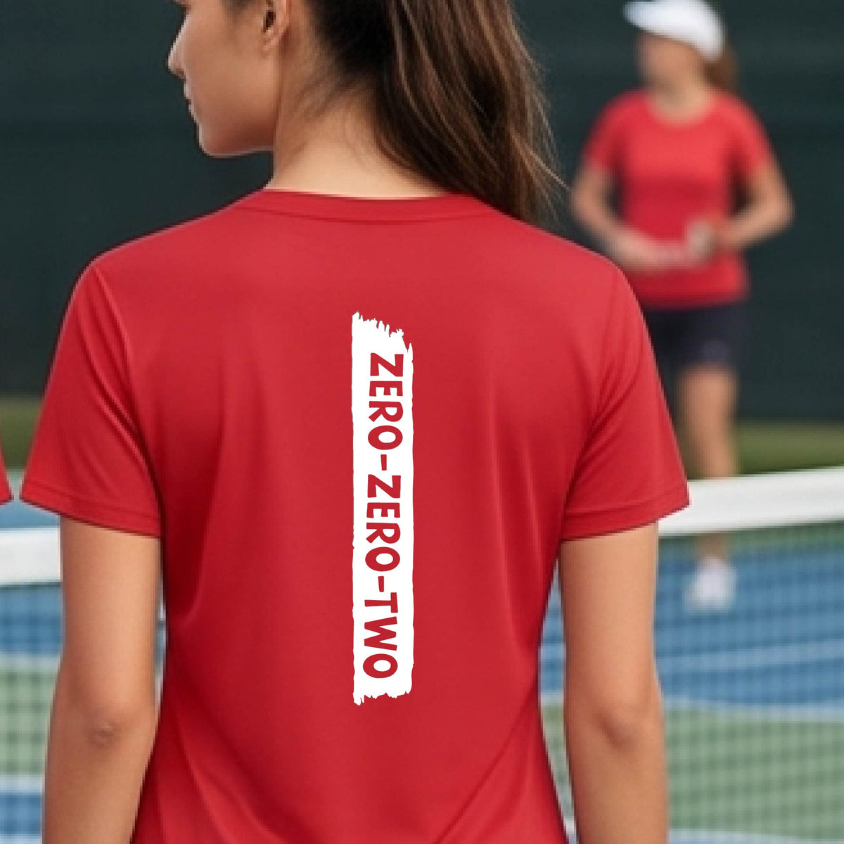 A close-up of a woman on a pickleball wearing a red short-sleeve v-neck t-shirt, with a white graphic of artistic banner with the cutout words of ‘ZERO ZERO TWO’, on the back of the shirt.