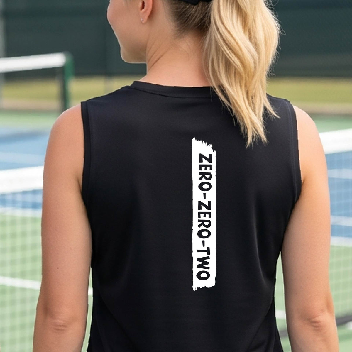 A close-up of a woman on a pickleball wearing a black sleeveless t-shirt, with a white graphic of artistic banner with the cutout words of ‘ZERO ZERO TWO’, on the back of the shirt.