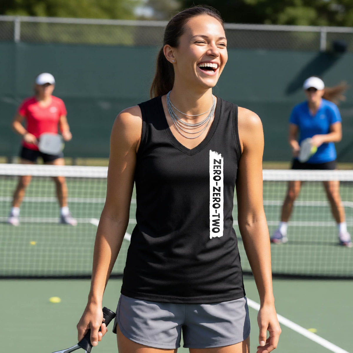 A close-up of a woman on a pickleball wearing a black sleeveless t-shirt, with a white graphic of artistic banner with the cutout words of ‘ZERO ZERO TWO’, on the front of the shirt.