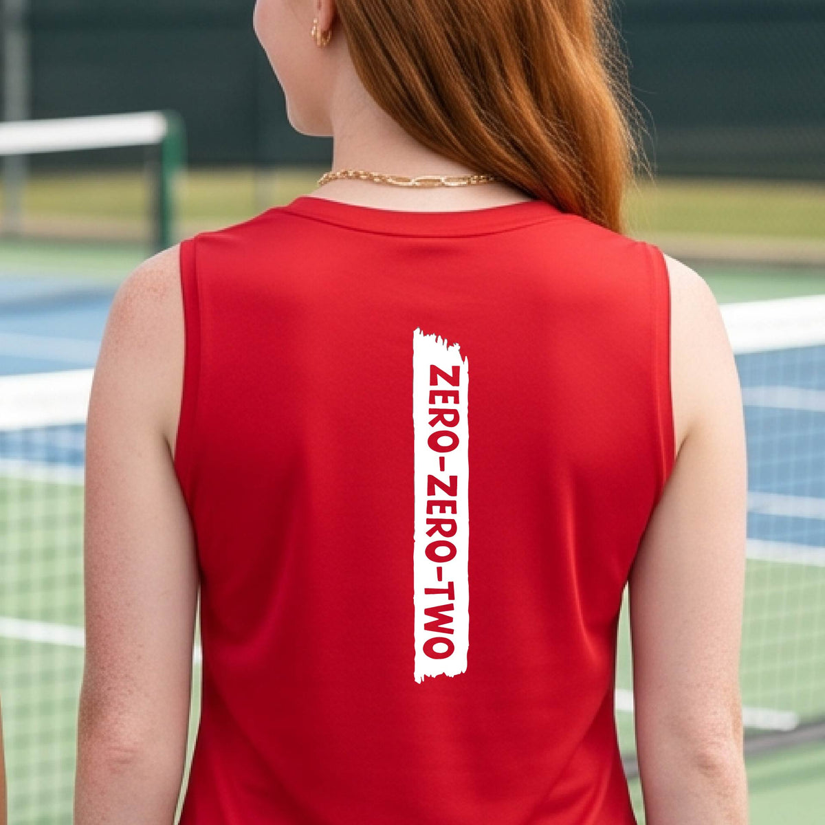 A close-up of a woman on a pickleball wearing a red sleeveless t-shirt, with a white graphic of artistic banner with the cutout words of ‘ZERO ZERO TWO’, on the back of the shirt.