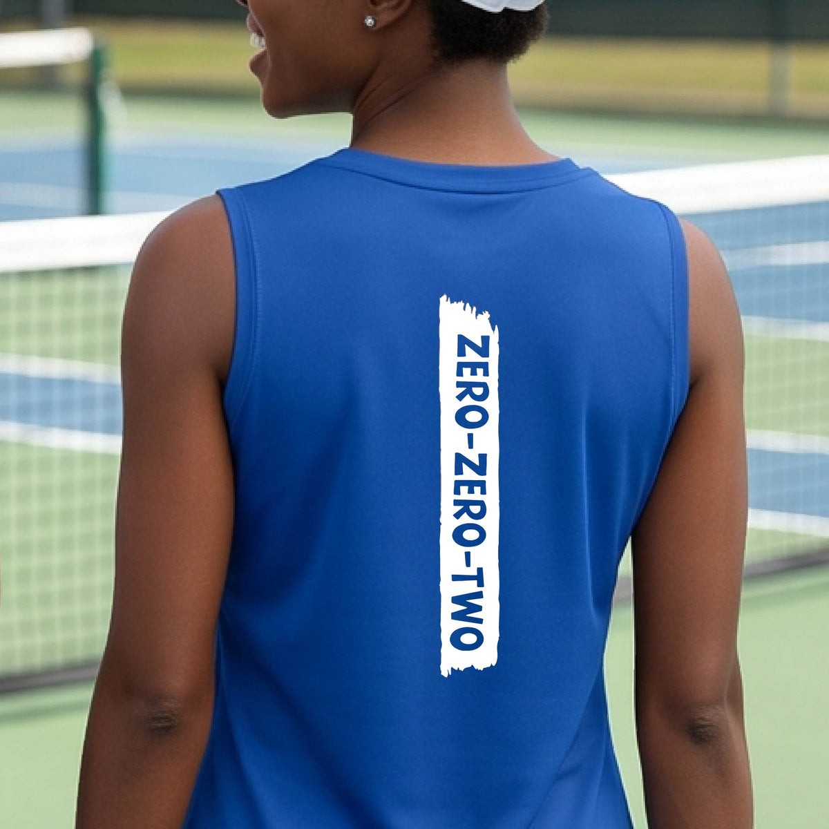 A close-up of a woman on a pickleball wearing a royal blue sleeveless t-shirt, with a white graphic of artistic banner with the cutout words of ‘ZERO ZERO TWO’, on the back of the shirt
