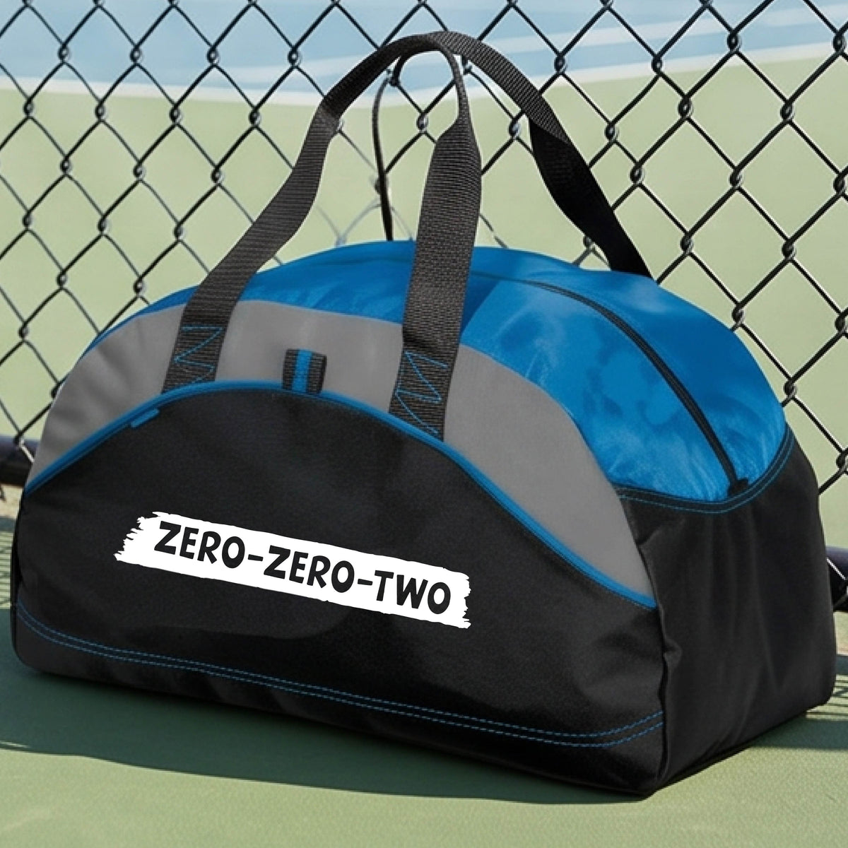 A close-up of a blue and black duffel bag with gray panels and a white graphic of artistic banner with the cutout words of ‘ZERO ZERO TWO’, on the front. 